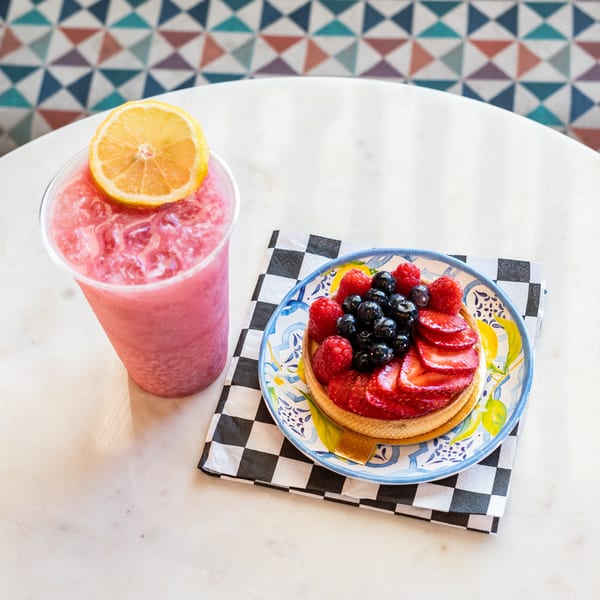 A berry tart on a decorative plate with a checkered napkin next to a pink drink with a lemon slice.
