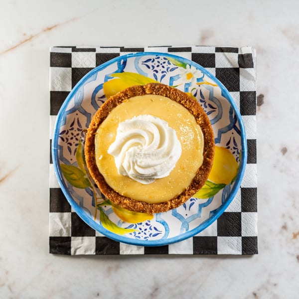 Overhead view of a key lime pie tart with whipped cream on a lemon-patterned plate and checkered napkin.