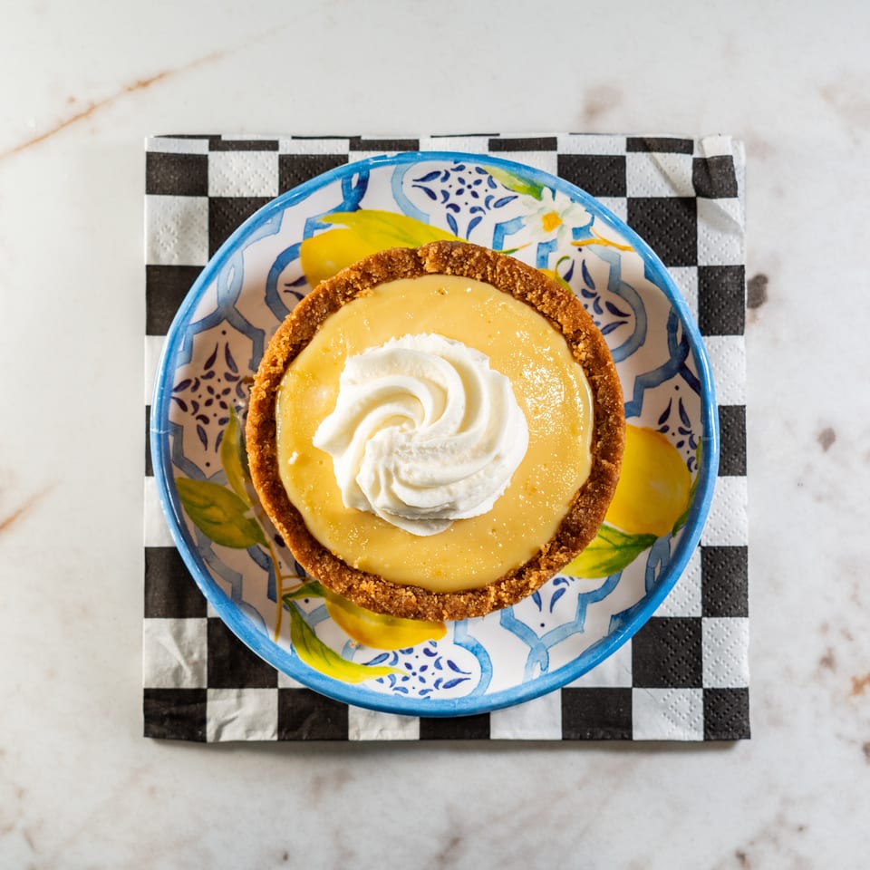 Overhead view of a key lime pie tart with whipped cream on a lemon-patterned plate and checkered napkin.