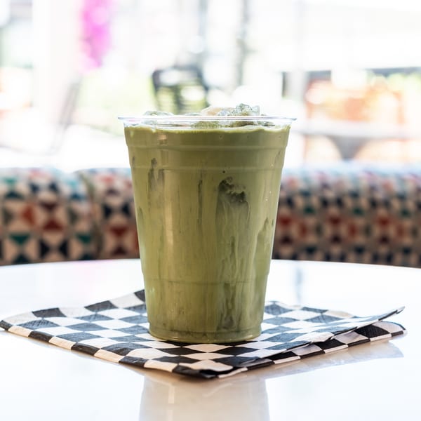 Iced green matcha latte in a clear plastic cup on a checkered napkin, on a white table.