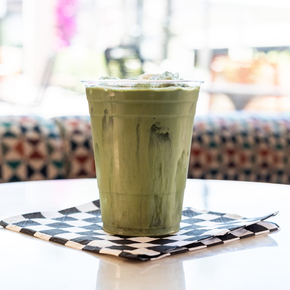 Iced green matcha latte in a clear plastic cup on a checkered napkin, on a white table.